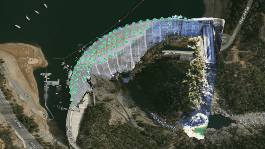 Aerial view of a dam with numerous green markers, enhanced by iTwin IoT Asset Analytics, along its curved top edge, surrounded by roads, water, and trees.