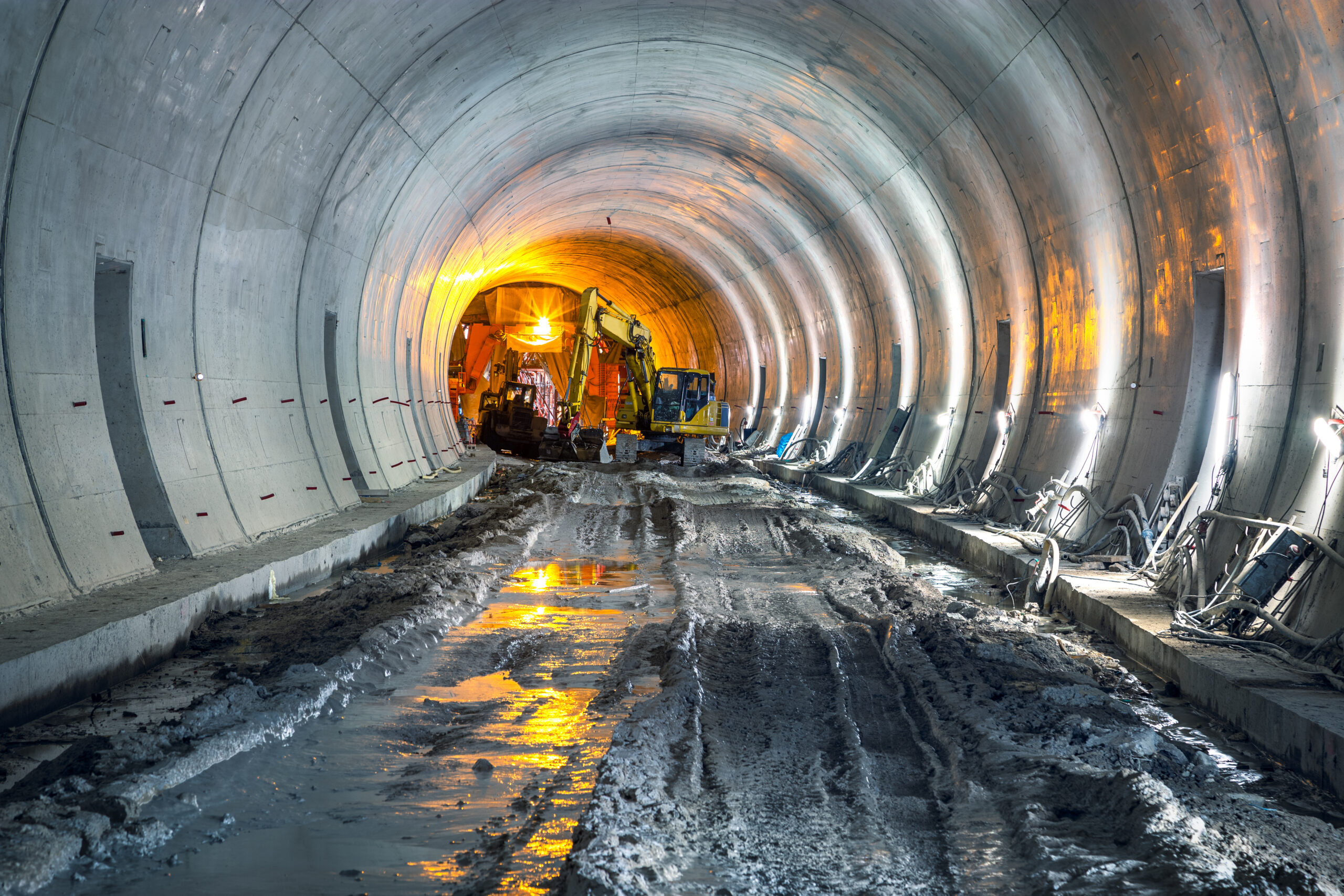 Construction equipment and workers are active inside a partially completed and illuminated tunnel with a muddy floor, focusing on geotechnical tunnel modelling.